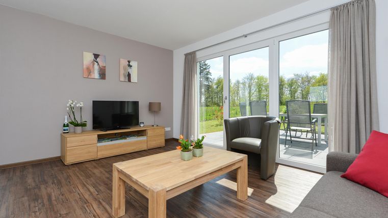 Ein modernes Wohnzimmer mit einem Holzschreibtisch, einem Fernseher und großen Fenstern. Der Raum ist hell und freundlich mit Blick auf einen Garten.