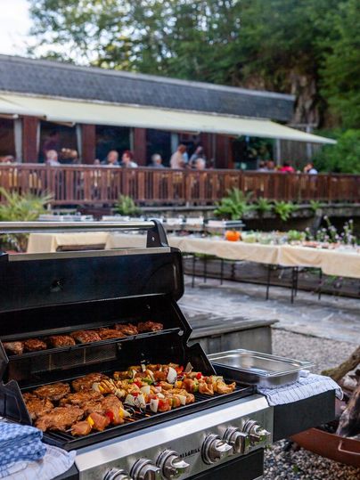 Ein Grill steht im Vordergrund, auf dem köstliches Fleisch und Gemüse zubereitet werden. Im Hintergrund sind Gäste in einem gemütlichen Restaurant zu sehen.