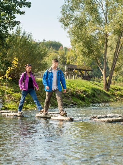 Zwei Kinder überqueren einen kleinen Fluss auf Steinen. Im Hintergrund sind Bäume und eine Sitzgelegenheit zu sehen.