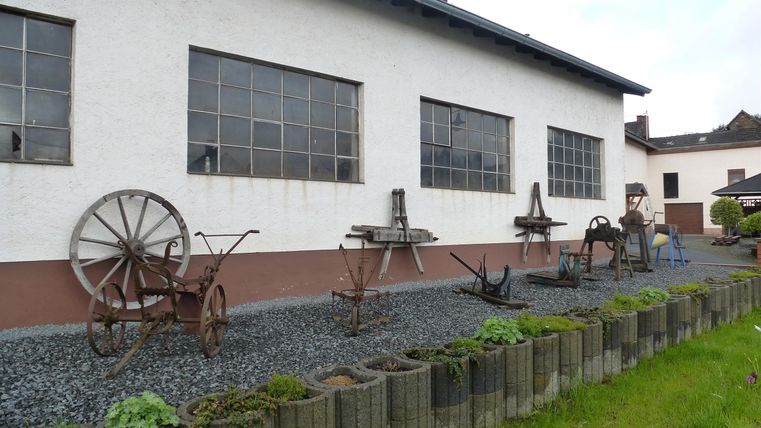 Eine Sammlung von historischen landwirtschaftlichen Geräten steht vor einem weißen Gebäude. Im Hintergrund sind große Fenster sichtbar, die einen Einblick in den Innenraum gewähren.