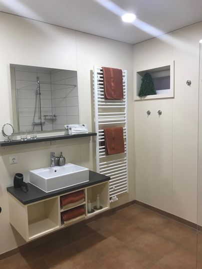 Ein modernes Badezimmer mit einem Waschbecken und einem Spiegel. Handtücher hängen an einem Handtuchheizkörper und der Boden ist braun gefliest.