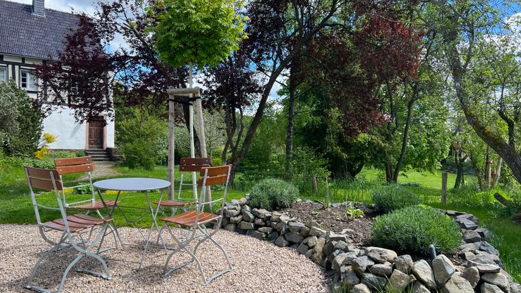 Ein gemütlicher Garten mit einem runden Tisch und Stühlen aus Metall. Im Hintergrund sind Bäume und eine grüne Wiese zu sehen.