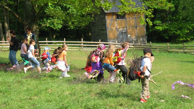 A group of children is playing outside and happily running across a meadow. In the background, a wooden building stands and trees surround the scene.