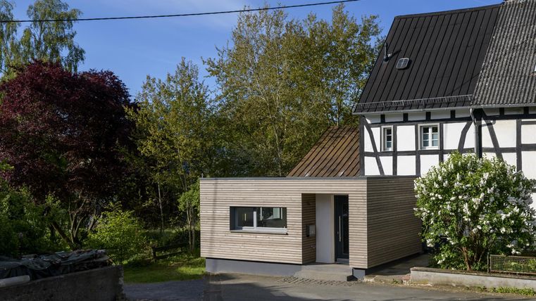 Ein modernes, helles Gebäude steht neben einem traditionellen, Fachwerkhaus. Umgeben von grünen Bäumen und Sträuchern vermittelt die Szene eine harmonische Atmosphäre.