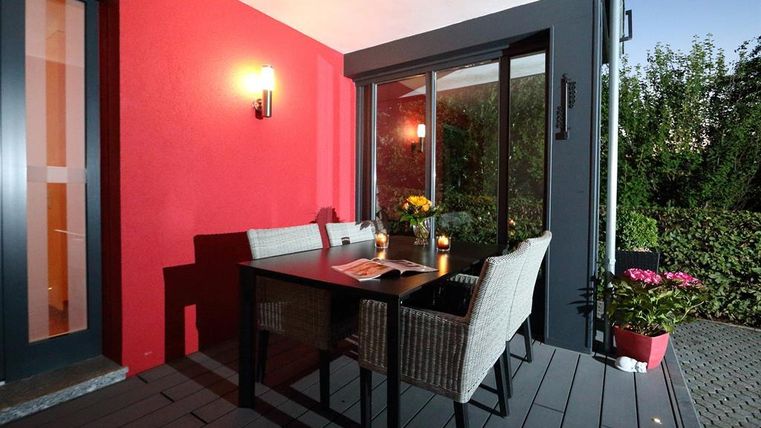 Een gezellige terras met een tafel en stoelen. Op de achtergrond zijn planten en een roodgeschilderde muur te zien.