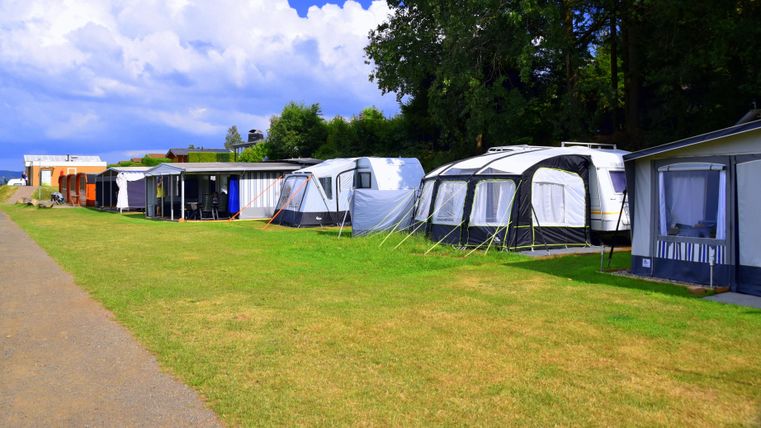 Wohnwagen mit Vorzelten auf einem grünen Campingplatz, umgeben von Bäumen und blauem Himmel.