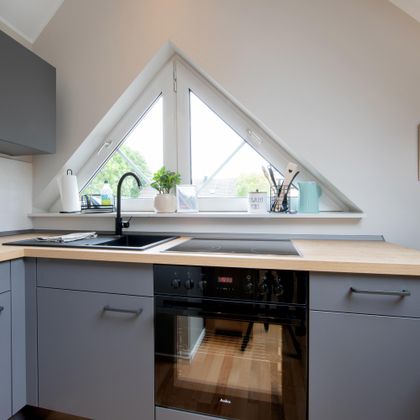 Een moderne keuken met grijze kasten en een houten werkblad. Grote ramen zorgen voor veel natuurlijk licht en een aangename sfeer.