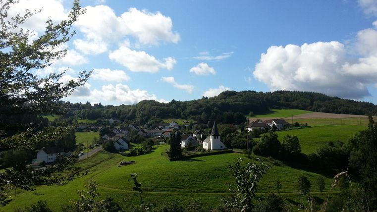 A picturesque landscape with a small village and a church. The sky is blue with some clouds and the surroundings are green and hilly.