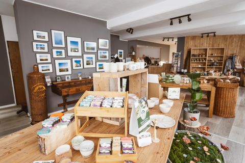 The image shows a store with handmade soaps, ceramic goods, and decorative items displayed on wooden tables and shelves. Framed photographs of landscapes hang on the wall.
