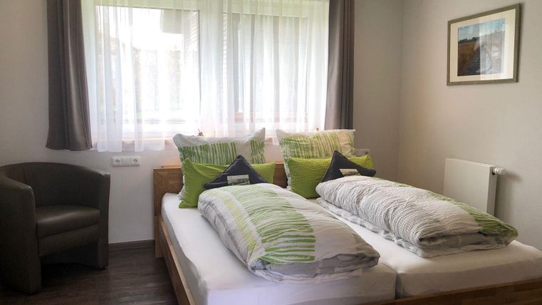 Ein helles Schlafzimmer mit zwei Betten und bunten Kissen. Eine gemütliche Sitzecke und große Fenster sorgen für eine einladende Atmosphäre.