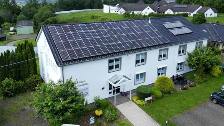 A modern building with solar panels on the roof. The entrance area is surrounded by plants and green spaces.