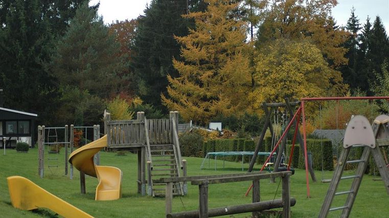 Ein Spielplatz mit Rutsche, Klettergerüst und Schaukel, umgeben von herbstlichen Bäumen.