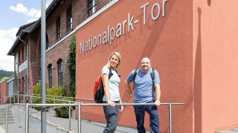 Zwei Personen stehen lächelnd vor dem Nationalpark-Tor. Im Hintergrund ist ein rotes Gebäude mit der Aufschrift "Nationalpark-Tor" zu sehen.