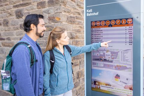 Een paar staat voor een perronbord en kijkt naar de informatie. De vrouw wijst met haar vinger naar de vertrektijden.
