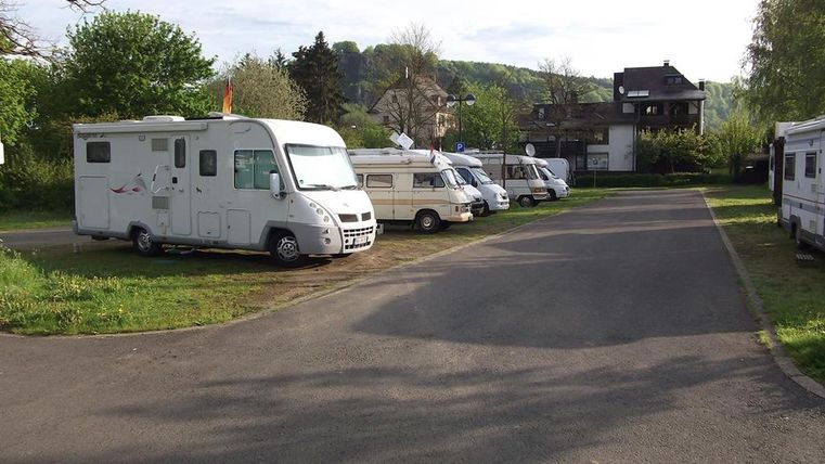Wohnmobile parken auf einem Stellplatz neben einer Straße, umgeben von Bäumen und Gebäuden im Hintergrund.