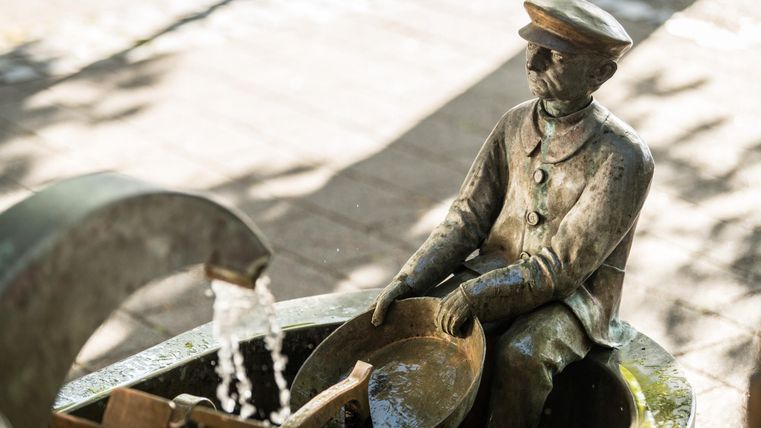 Eine Statue eines Mannes im Uniform mit einer Schaufel in den Händen. Wasser fließt aus einer Quelle in der Nähe.
