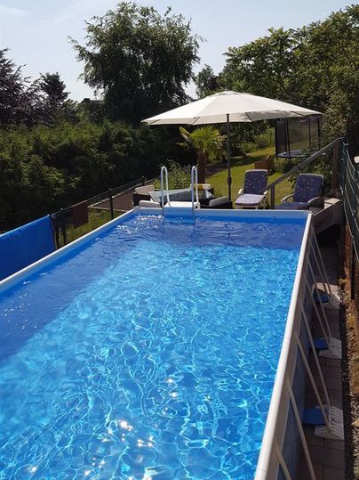 Ein blauer Swimmingpool mit klarem Wasser und zwei Liegen. Im Hintergrund sind Bäume und ein Sonnenschirm zu sehen.