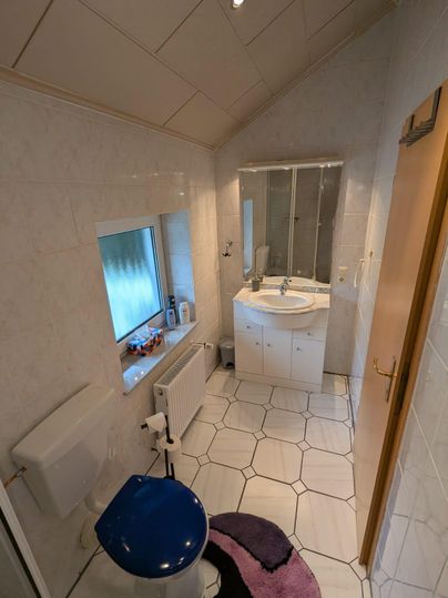 A bathroom with a toilet, sink, and mirror. The walls are light-colored tiled, and there is a window that lets in natural light.