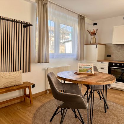 Eine helle und moderne Küche mit einem runden Holztisch und bequemen Stühlen. An der Wand sind dekorative Elemente und große Fenster, die natürlichen Licht hereinlassen.