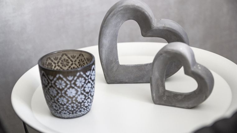 A stylish decorative arrangement with two heart-shaped sculptures made of concrete and a beautiful, patterned pot. The objects are on a white table and create a modern atmosphere.