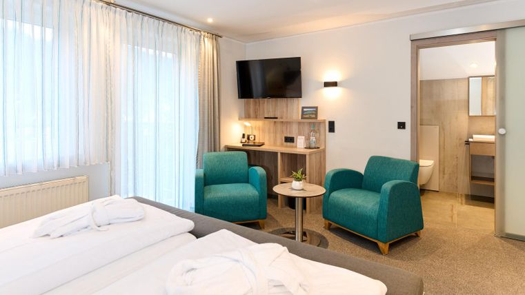 Een modern hotelkamer met twee bedden, een zithoek en een televisie. De lichte gordijnen en de groene stoelen zorgen voor een vriendelijke sfeer.