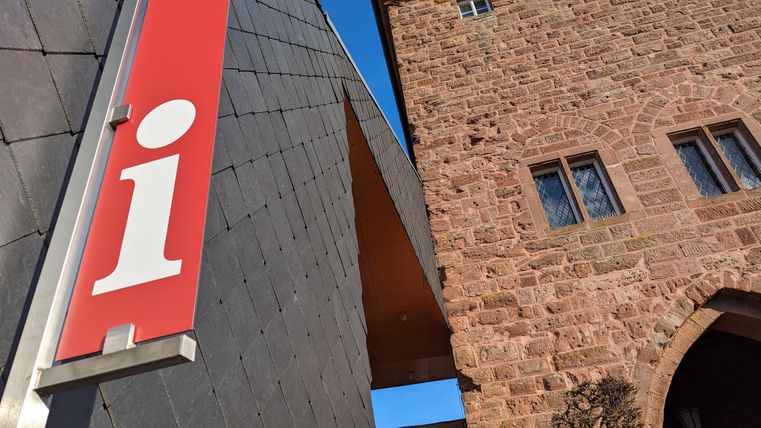 Ein Informationsschild in Rot mit einem großen „i“ steht im Vordergrund. Im Hintergrund ist ein historisches Gebäude aus Stein zu sehen.