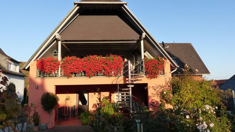 A beautiful house with a red flower box and a balcony. In the foreground, green plants and a blue sky are visible.