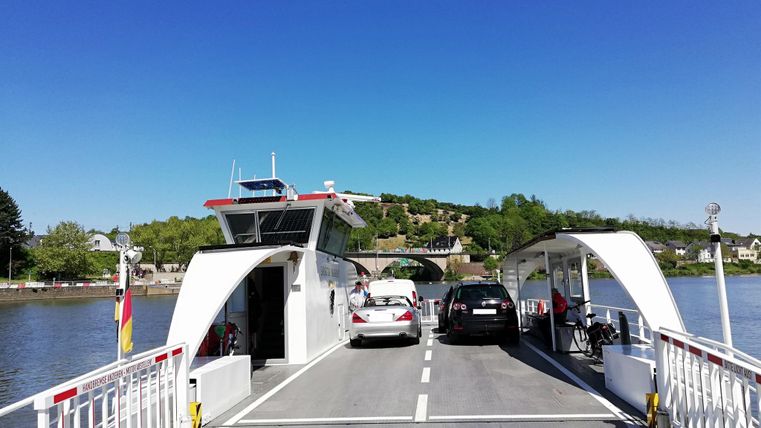 A car ferry on a calm river with green hills in the background. The sky is clear and blue.