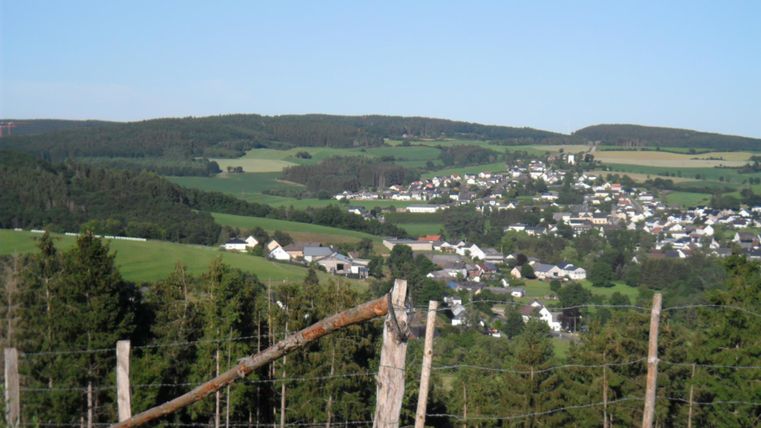 Eine malerische Landschaft mit Hügeln und einem kleinen Dorf im Tal. Im Vordergrund sind Weiden und Zaunpfähle sichtbar.