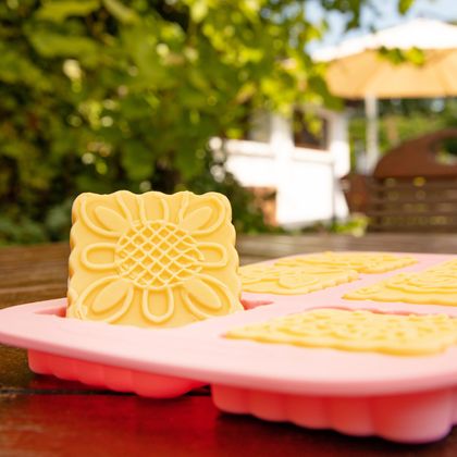 Eine Silikonform mit gelbem, blumenförmigem Gebäck steht auf einem Tisch. Im Hintergrund sind grüne Blätter und ein sonniger Garten zu sehen.