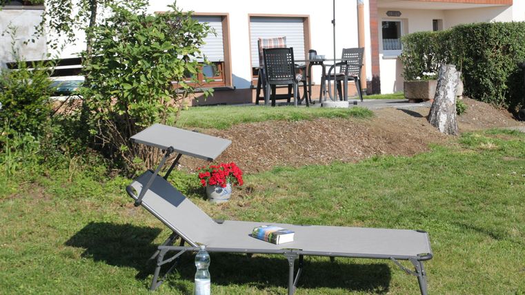 Een ligstoel staat op een groene wei, omringd door planten. Op de achtergrond zijn een tafel en stoelen onder een parasol te zien.