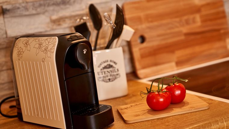 A modern coffee machine is standing on a wooden table. Next to it are fresh tomatoes and kitchen utensils.