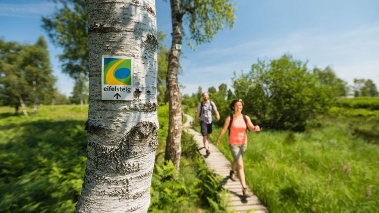 A woman and a man are hiking on a wooden path through a green landscape. A trail marker sign for the Eifelsteig is visible on a tree.