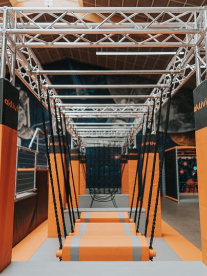 Ein moderner Abenteuerpark mit hängenden Seilen und gepolsterten Plattformen. Die orange und graue Ausstattung lädt zum Spielen und Klettern ein.