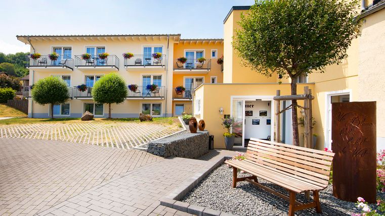 A cozy entrance area with a light yellow building. A bench and large, green trees create an inviting atmosphere.
