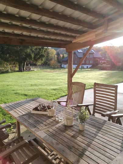 Ein gemütlicher Außenbereich mit einem Holztisch und Stühlen. Im Hintergrund ist eine grüne Wiese und ein sonniger Himmel sichtbar.