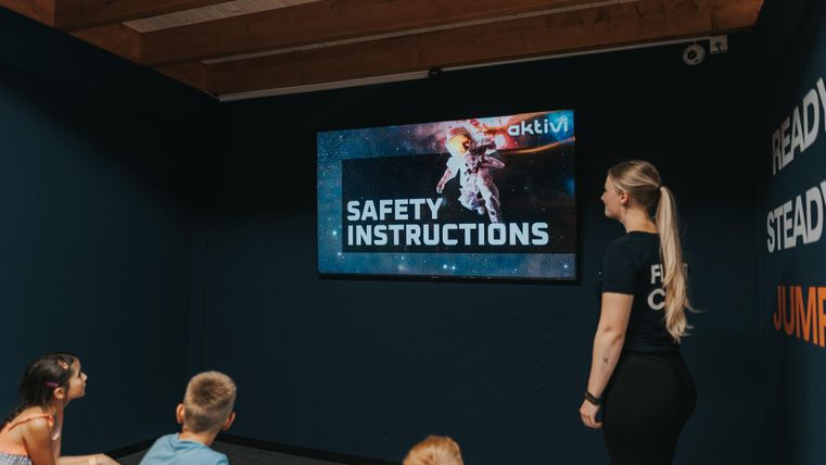 Eine Gruppe von Kindern hört einer Anleitung zu Sicherheitshinweisen zu, während sie in einem Raum sitzen. Eine Lehrerin steht vor einem Bildschirm, auf dem die Sicherheitsanweisungen angezeigt werden.