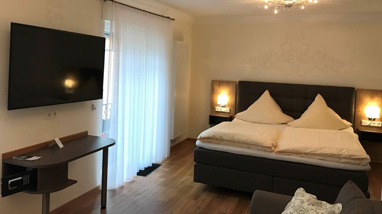 Ein modernes Schlafzimmer mit einem Doppelbett, einem großen Fernseher und einem gemütlichen Sessel. Das Licht ist freundlich, und das Zimmer hat große Fenster.