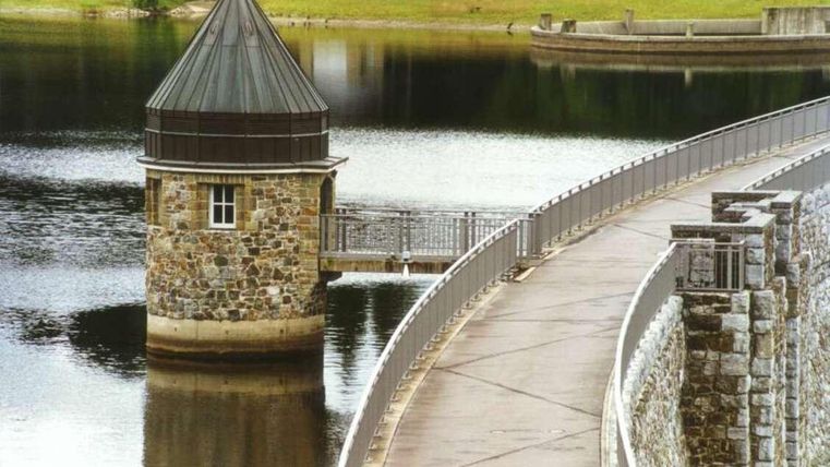 Ein schöner Wasserturm aus Steinen am Ufer eines ruhigen Sees. Im Hintergrund sieht man eine wellenförmige Uferpromenade.