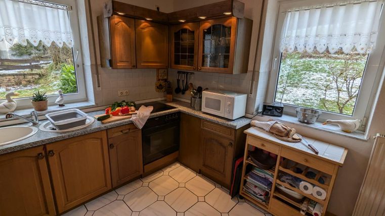 A cozy kitchen with wooden cabinets and light tiles. Next to the window, there is a small table and various kitchen appliances.