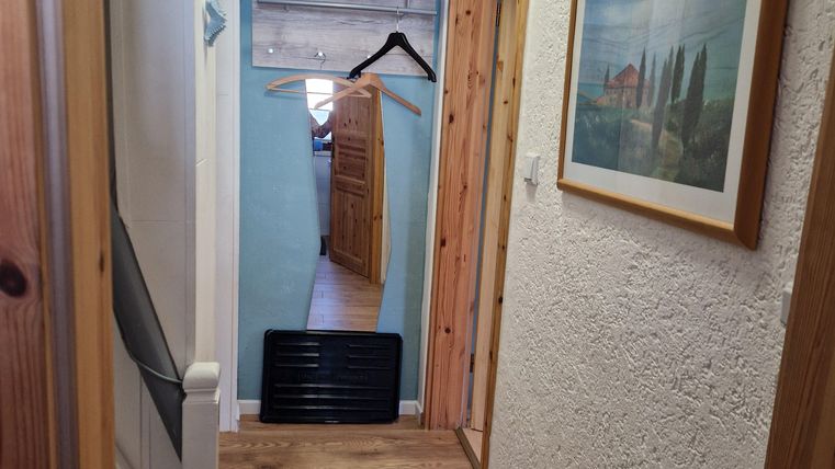 Een lichte gang met houten vloer en witte muren. Op de achtergrond zijn een fotolijst en een kledingstandaard te zien.