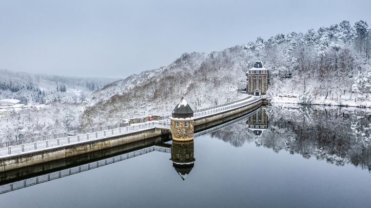 Eine winterliche Landschaft mit einer schneebedeckten Staumauer und stillem Wasser. Bäume im Hintergrund sind ebenfalls mit Schnee bedeckt, was eine ruhige Atmosphäre schafft.