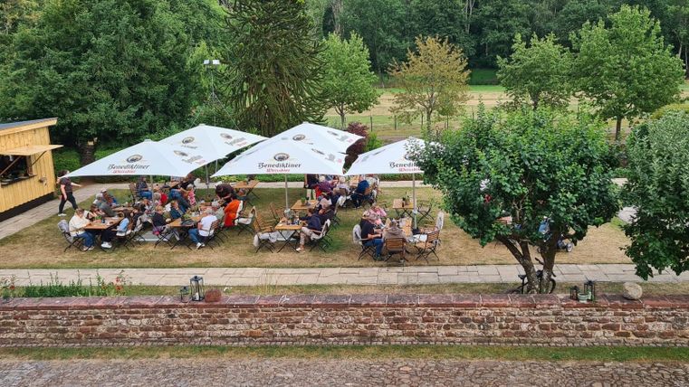 Eine gemütliche Terrasse mit vielen Gästen und Sonnenschirmen. Umgeben von Bäumen und einer grünen Wiese.