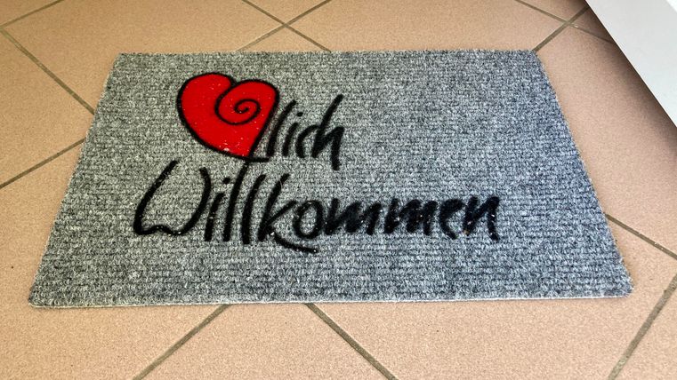 Ein grauer Eingangsteppich mit der Aufschrift "Willkommen" und einem roten Herz. Er vermittelt eine freundliche Atmosphäre für Gäste.
