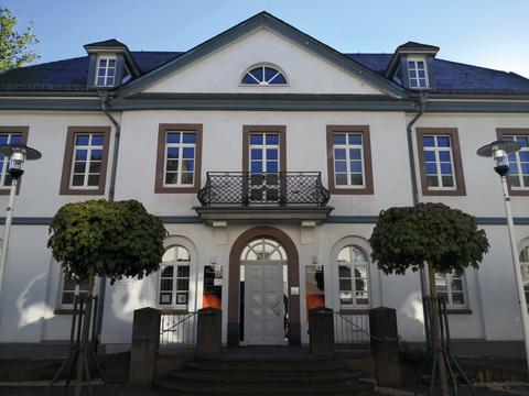 Ein elegantes Gebäude mit weißen Wänden und großen Fenstern. Im Vordergrund stehen zwei runde, grüne Bäume.