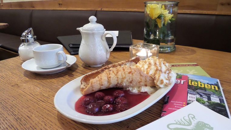 Ein leckerer Crepe mit Kirschen auf einem weißen Teller. Im Hintergrund sind eine Teekanne und eine Blume in einer Vase zu sehen.