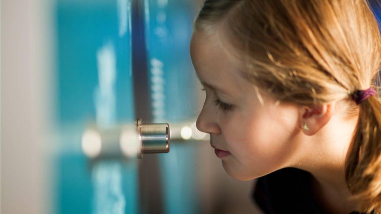Ein Mädchen schaut neugierig durch ein kleines Fenster an einer Wand. Im Hintergrund sind unscharfe, helle Farben zu sehen.