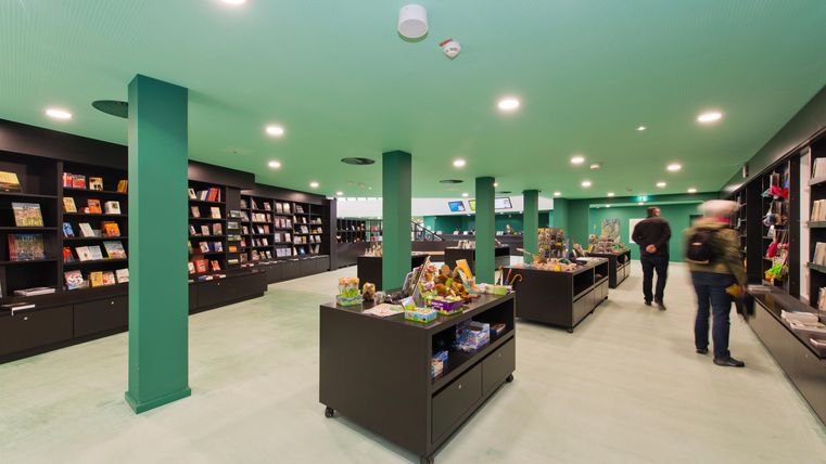 A modern bookstore with green walls and elegant shelves. Customers browse between the shelves and look at various books.