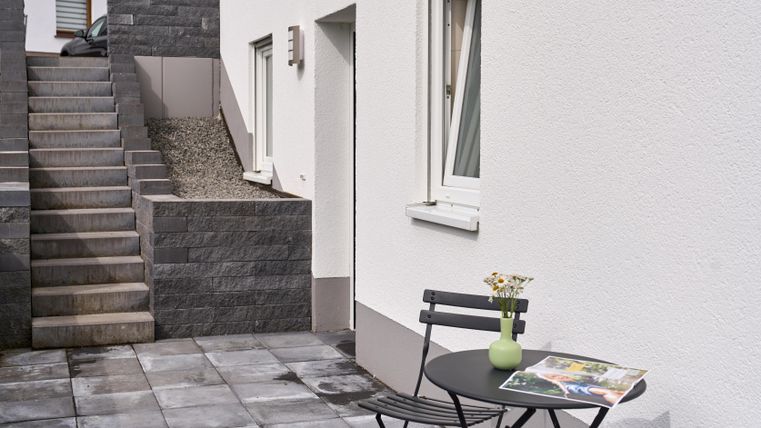 Ein kleiner Außenbereich mit einer schwarzen Tisch- und Stuhlgruppe. Eine Treppe führt zu einem höher gelegenen Bereich, während ein Blumenstrauß auf dem Tisch steht.