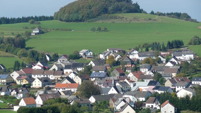 Een schilderachtig landschap met een klein dorp, omringd door groene weilanden en heuvels. De huizen zijn kleurrijk en goed gerangschikt, wat een idyllische sfeer creëert.
